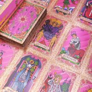 May include: A pink tarot card deck with a matching box. The cards feature illustrations and titles like "The Lovers" and "Strength." The deck is set against a pink floral background, creating a visually appealing aesthetic.