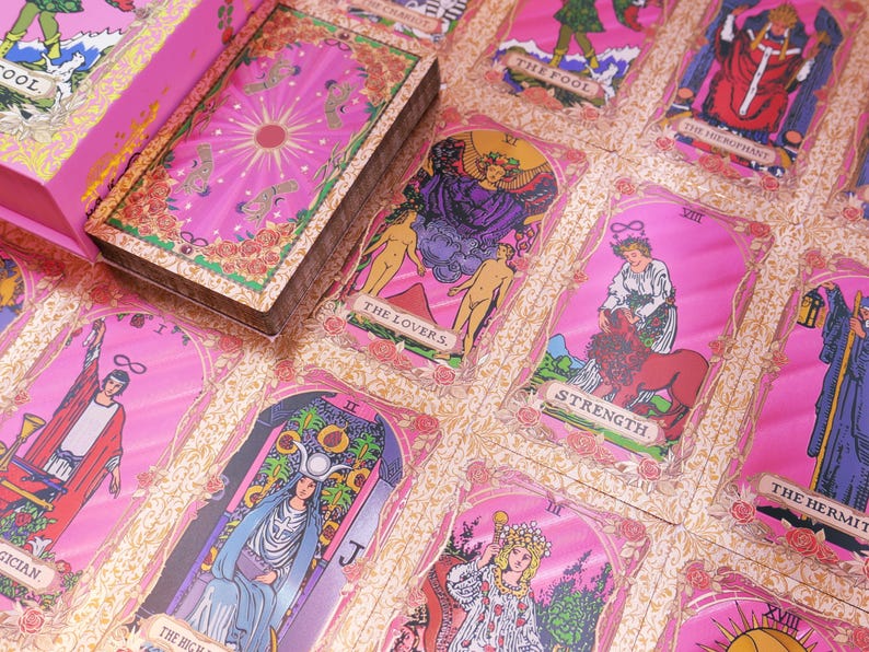 May include: A pink tarot card deck with a matching box. The cards feature illustrations and titles like "The Lovers" and "Strength." The deck is set against a pink floral background, creating a visually appealing aesthetic.