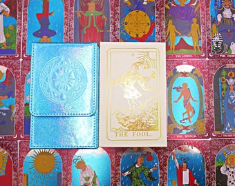 May include: A rectangular light blue iridescent card case with a circular embossed design next to a white tarot deck box featuring a gold foil illustration of The Fool and the text THE FOOL. The items are placed over a background of tarot cards.