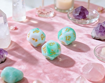 May include: Three multi-faceted dice with a white and green marbled finish and gold-colored engravings. One die features zodiac symbols, while the other two display numbers. The dice are arranged on a soft pink surface surrounded by various crystals.