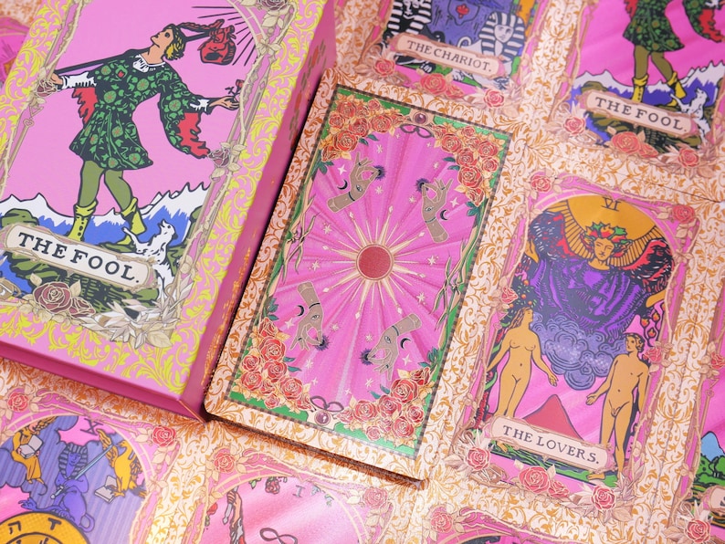 May include: A pink tarot card deck and individual cards featuring illustrations and text. The box and cards display images of figures, symbols, and text, including "THE FOOL" and "THE LOVERS." The cards have a pink background with floral and celestial designs.