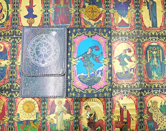 May include: A rectangular deck box with a holographic finish and a circular geometric emblem, placed next to a tarot card box featuring a colorful illustration of The Fool, surrounded by a background of various tarot cards.