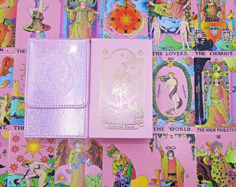 May include: A light pink tarot deck box with a glittery finish and a matching light pink tarot card box featuring gold foil illustrations and the text Gold foil Tarot, displayed against a background of various colourful tarot cards.
