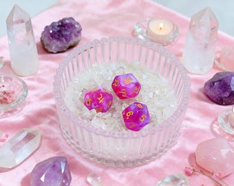 May include: Three magenta and white marbled polyhedral dice with gold-colored zodiac symbols and numbers, resting inside a clear, ribbed glass bowl filled with small, translucent white crystal chips, surrounded by various quartz and amethyst stones.