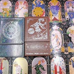 May include: A black holographic card case with a circular silver emblem next to a brown box labelled Gold foil Tarot 78 latest Waite Taro decks, featuring a silver illustration of a figure and the word GENUINE on the front cover.