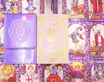 May include: A rectangular purple iridescent deck box with a circular embossed design next to a light pink tarot card deck box featuring a gold foil illustration of a figure standing in an archway, set against a background of various tarot cards.