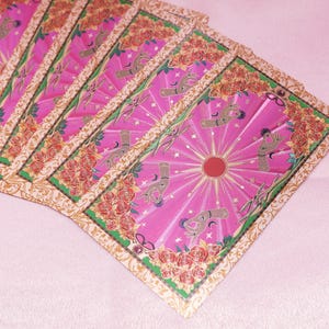 May include: A fan of tarot cards with a pink background. The cards feature a pink and gold design with a central sunburst, hands, and floral borders. The cards are arranged in a fanned-out pattern.