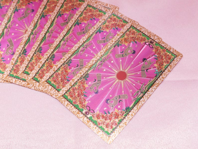 May include: A fan of tarot cards with a pink background. The cards feature a pink and gold design with a central sunburst, hands, and floral borders. The cards are arranged in a fanned-out pattern.