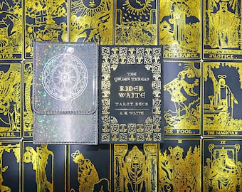 May include: A black tarot deck box with a holographic circular emblem and a matching black tarot card box featuring gold foil text that reads The Golden Thread Rider Waite Tarot Deck A. E. Waite, set against a background of gold tarot cards.