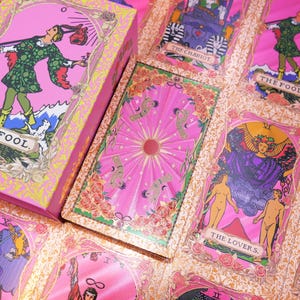 May include: A pink tarot card deck and box set. The cards feature illustrations with pink, green, and gold accents. The box and cards display the text "THE FOOL" and other card names. The deck is decorated with floral and celestial designs.