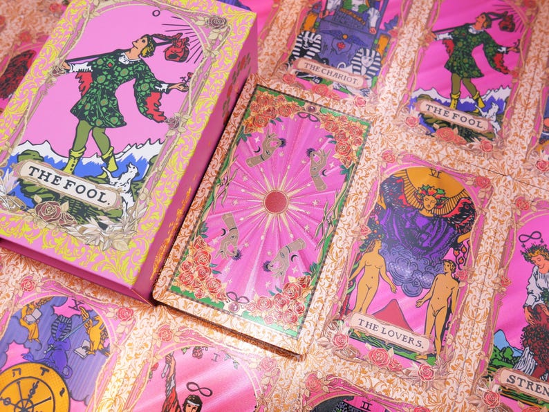 May include: A pink tarot card deck and box set. The cards feature illustrations with pink, green, and gold accents. The box and cards display the text "THE FOOL" and other card names. The deck is decorated with floral and celestial designs.