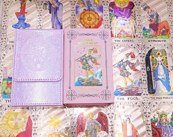 Puede incluir: Una caja de cartas de tarot de color morado claro con purpurina junto a una caja de cartas de tarot de color morado claro con una ilustración en lámina dorada de The Fool y el texto Gold Foil Tarot y The Fool, sobre un fondo de cartas.