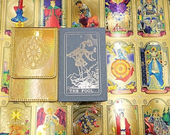 May include: A gold holographic tarot card pouch with a circular sun emblem next to a dark grey tarot deck box featuring a gold-foiled illustration of The Fool card, all displayed against a background of various gold-accented tarot cards.
