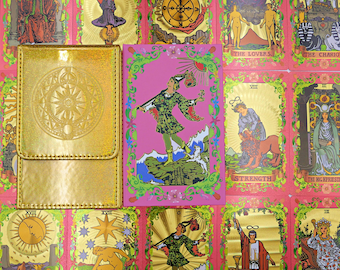 May include: A gold metallic rectangular card case with a circular embossed sun design sits next to a pink tarot card featuring a figure in green clothing walking on a cliff, surrounded by a background of various gold-foiled tarot cards.