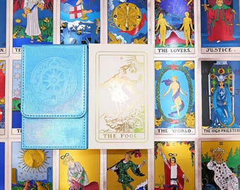 May include: A light blue iridescent deck box with a circular emblem embossed on the flap, positioned next to a white tarot card box featuring a gold foil illustration of The Fool and the text THE FOOL, set against a background of tarot cards.