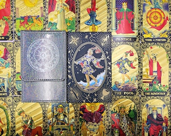 May include: A rectangular deck box with a holographic geometric circle design sits beside a matching tarot card box featuring a central illustration of a figure walking, surrounded by gold celestial motifs on a dark background.