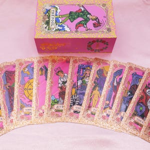 May include: A pink tarot card deck with a matching box. The cards are fanned out, displaying various illustrations with pink and gold borders. The box features an image of a person and the words "THE FOOL".