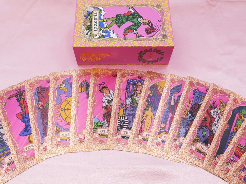 May include: A pink tarot card deck with a matching box. The cards are fanned out, displaying various illustrations with pink and gold borders. The box features an image of a person and the words "THE FOOL".