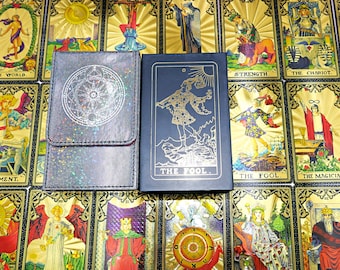 May include: A rectangular black tarot card box with a gold foil illustration of The Fool and the text THE FOOL, positioned next to a dark, iridescent, textured card pouch featuring a circular silver celestial design on the flap.