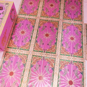 May include: A pink tarot card deck with a matching box. The cards are pink with a central sunburst design and floral borders. The box features an illustration of a figure in green and red clothing.