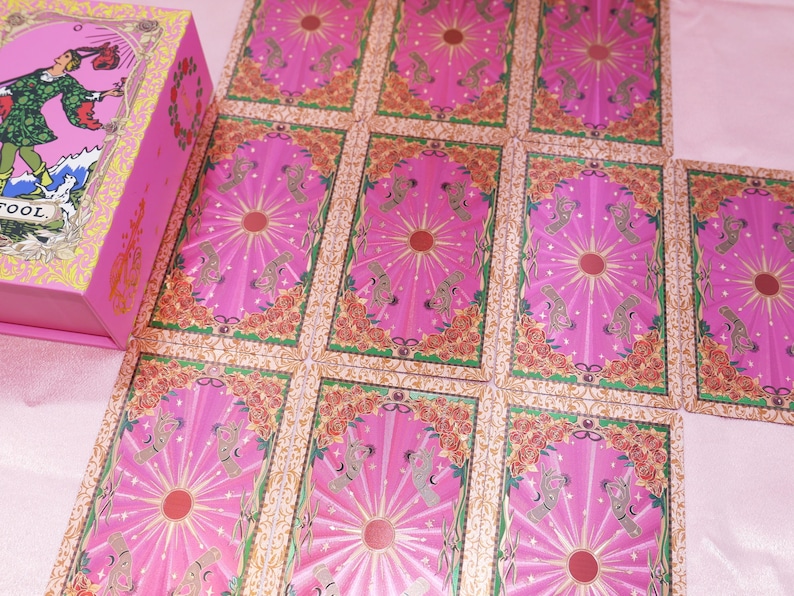 May include: A pink tarot card deck with a matching box. The cards are pink with a central sunburst design and floral borders. The box features an illustration of a figure in green and red clothing.