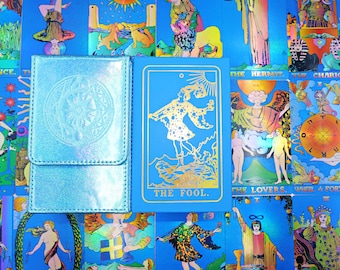 May include: A light blue tarot deck box with a circular embossed design next to a blue tarot card featuring a gold foil illustration of The Fool, set against a background of various colorful tarot cards with holographic accents.