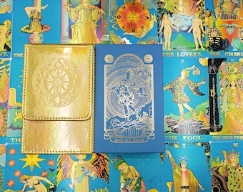 Puede incluir: Un estuche para cartas rectangular dorado iridiscente con un diseño circular en relieve junto a una caja de cartas azul con una ilustración en lámina dorada de una figura sobre un acantilado y el texto Gold Foil Tarot sobre un fondo de cartas.