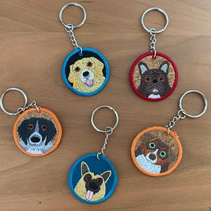 May include: Five keychains with embroidered dog portraits. Each keychain features a different breed of dog, including a golden retriever, a French bulldog, a black and white dog, a tri-colored dog, and a brown and white dog. The keychains are made with colorful fabric and have silver key rings.