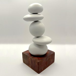 May include: A sculpture consisting of five smooth, light grey, rounded stones stacked vertically in a balanced column. The stack of stones is mounted on a square, dark reddish-brown wooden base with a visible wood grain texture and finish.