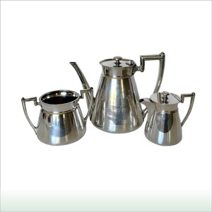 Puede incluir: Un juego de té plateado, que incluye una tetera, una jarra de crema y un azucarero. La tetera tiene una forma cónica alta con tapa y asa curva. La jarra de crema y el azucarero tienen asas a juego y un acabado pulido. El juego se muestra sobre fondo blanco.
