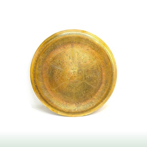May include: A round, gold-coloured metal tray with intricate engraved designs. The tray has a raised rim and a central design with a starburst pattern.