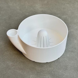 Op de afbeelding: Een witte citruspers met een ingebouwde ruimer en een kleine tuit. De pers heeft een ronde komvorm met een handvat en is gemaakt van een glad, stevig materiaal. De ruimer heeft verticale groeven voor efficiënt persen.