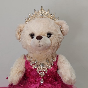 May include: A plush teddy bear wearing a pink dress with gold glitter, a gold crown with rhinestones, and a gold necklace with rhinestones.