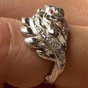 Könnte beinhalten: Ein silberner Ring mit Schlangenmotiv. Der Schlangenkopf hat ein rotes Edelsteinauge, und der Körper ist mit kleinen, klaren Edelsteinen besetzt. Der Ring hat ein detailliertes, strukturiertes Aussehen.