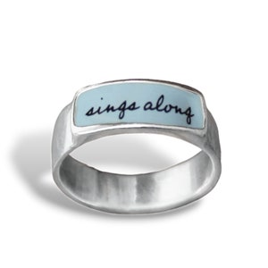 May include: A silver ring with a light blue rectangular inlay that says "sings along" in black script.