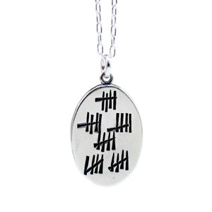 Oval shaped pendant with 30 hash marks crossed off in groups of five like on a prison wall. Reverse side has the words "who's counting" in center.