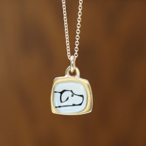 May include: A silver necklace with a pendant featuring a dog silhouette. The pendant has a gold-colored frame around a light blue square with a black dog outline. The chain is delicate and hangs against a brown background.