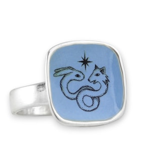May include: A silver ring with a square, light blue face. The face features a black illustration of a rabbit and a fox intertwined, with a star above them. The ring has a simple, elegant design.