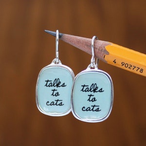 May include: A pair of silver dangle earrings with light blue rectangular charms that say "talks to cats" in black script.