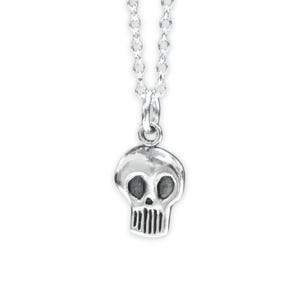 May include: A silver-toned necklace featuring a small, stylized skull pendant suspended from a delicate chain. The skull charm has dark, recessed eye sockets and vertical lines representing teeth, crafted with a polished metallic finish throughout.