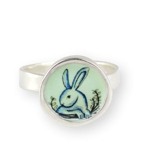 May include: A silver ring with a round, light green cameo featuring a blue and white rabbit illustration.