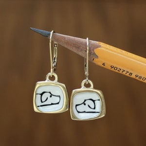 May include: A pair of gold-tone earrings with a white square pendant featuring a black silhouette of a dog's head.