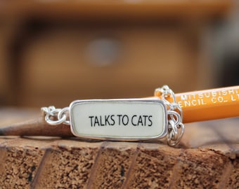 May include: A silver charm bracelet with a white rectangular charm that says "TALKS TO CATS" on it. The charm is attached to a silver chain with a pencil resting on top of it.