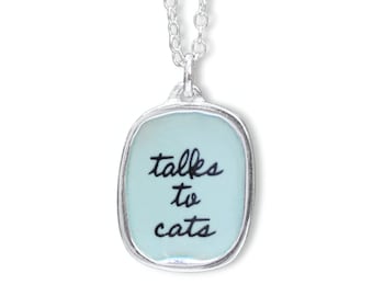 May include: A silver necklace with a light blue rectangular pendant that says "talks to cats" in black cursive writing.