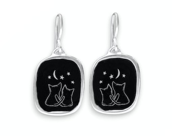 May include: Silver dangle earrings with a black enamel rectangular charm featuring two cats silhouetted against a crescent moon and stars.