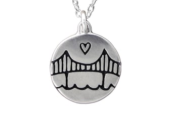 May include: A circular silver pendant necklace featuring an engraved bridge design with a small heart symbol above the bridge structure. The pendant hangs from a delicate silver chain, showcasing a matte metallic finish and dark etched lines.