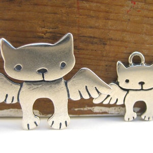 Puede incluir: Dos encantos de plata en forma de gatos con alas. El encanto m&aacute;s grande est&aacute; de pie sobre sus pies, mientras que el encanto m&aacute;s peque&ntilde;o est&aacute; colgando de un bucle.