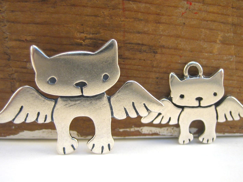 Puede incluir: Dos encantos de plata en forma de gatos con alas. El encanto m&aacute;s grande est&aacute; de pie sobre sus pies, mientras que el encanto m&aacute;s peque&ntilde;o est&aacute; colgando de un bucle.