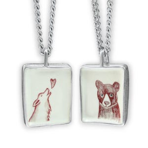 May include: Two silver necklaces with square pendants. The pendant on the left features a red wolf howling at a red heart. The pendant on the right features a red bear with a white nose.
