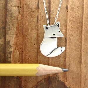 May include: A silver fox pendant necklace on a silver chain. The fox is sitting with its tail curled around its body. The necklace is on a wooden surface.